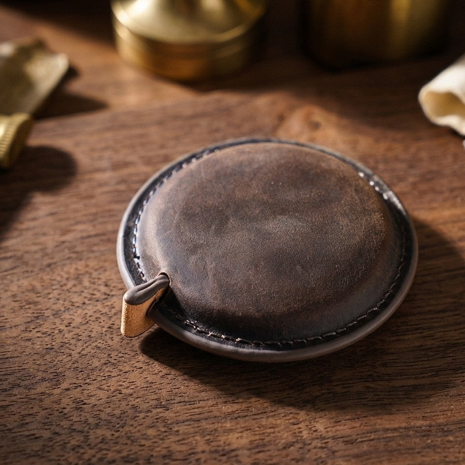 A round dark brown leather coin purse or small wallet with a stitched edge and a small leather tab, resting on a wooden tabletop.