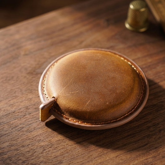 A small oval brown leather accessory with a stitched edge and a small tab, photographed on a wooden tabletop with a blurred brass object in the background.