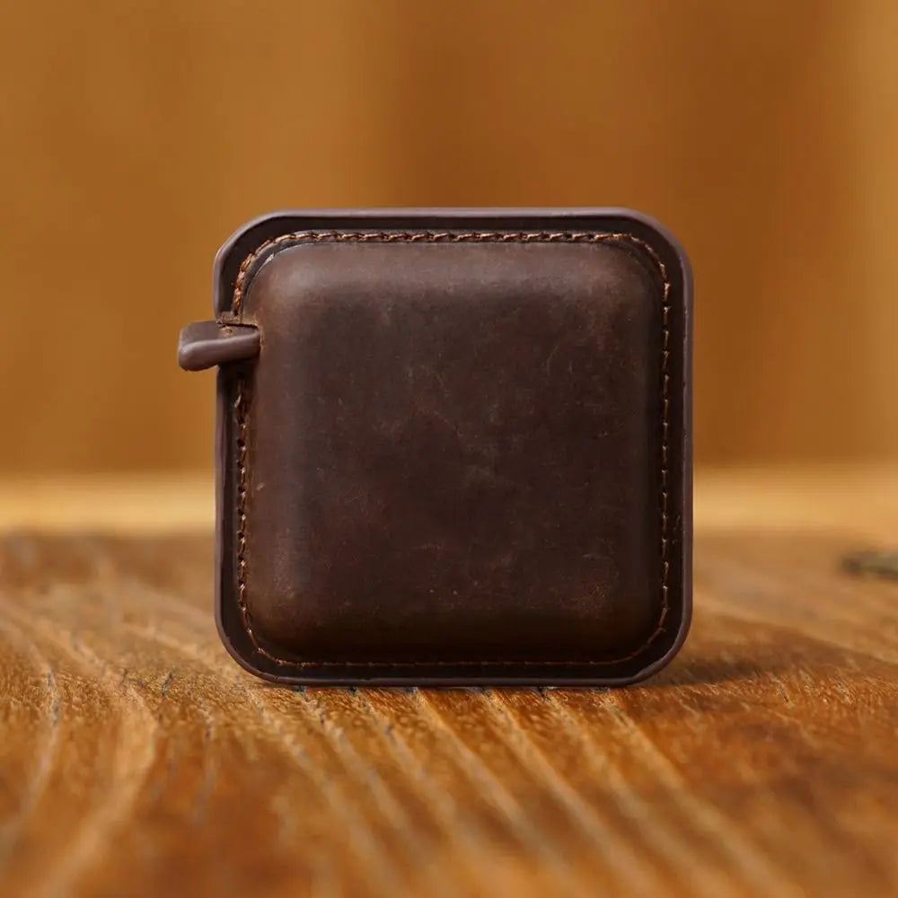 A square dark brown leather accessory with rounded corners, stitched edges, and a small loop tab on the left side, standing upright on a wooden surface against a warm blurred background.