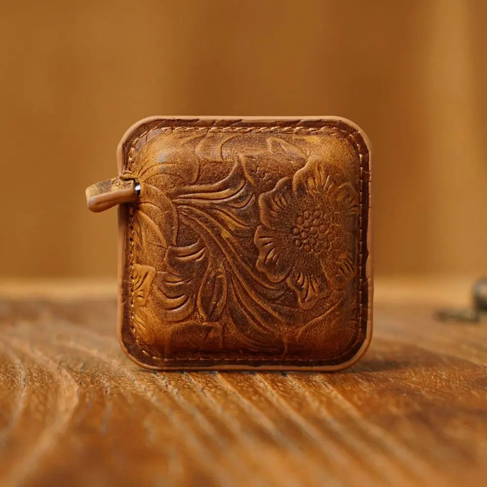 A small square brown leather pouch with an embossed floral pattern and a zipper pull, resting on a wooden surface with a warm blurred background.